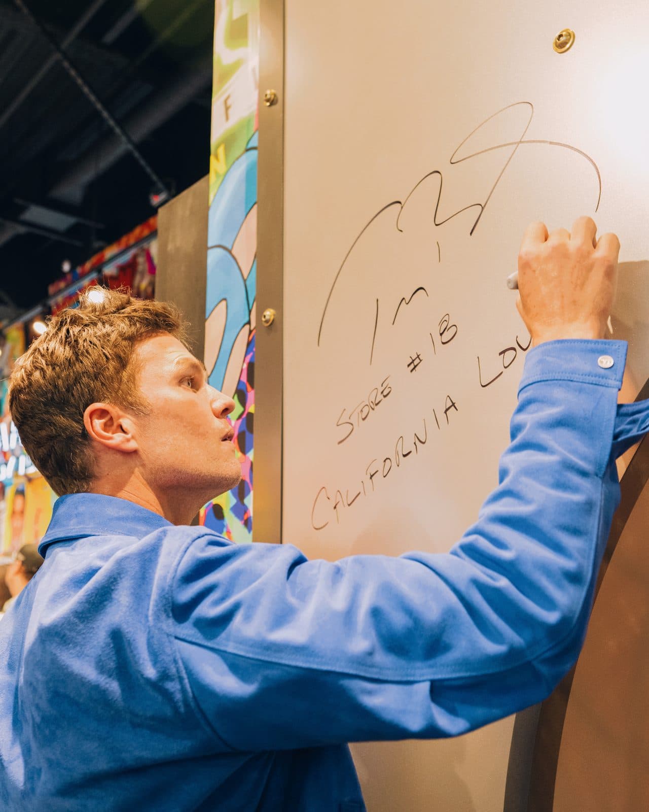 Tom Brady signs his name on a white wall panel inside the store's vault