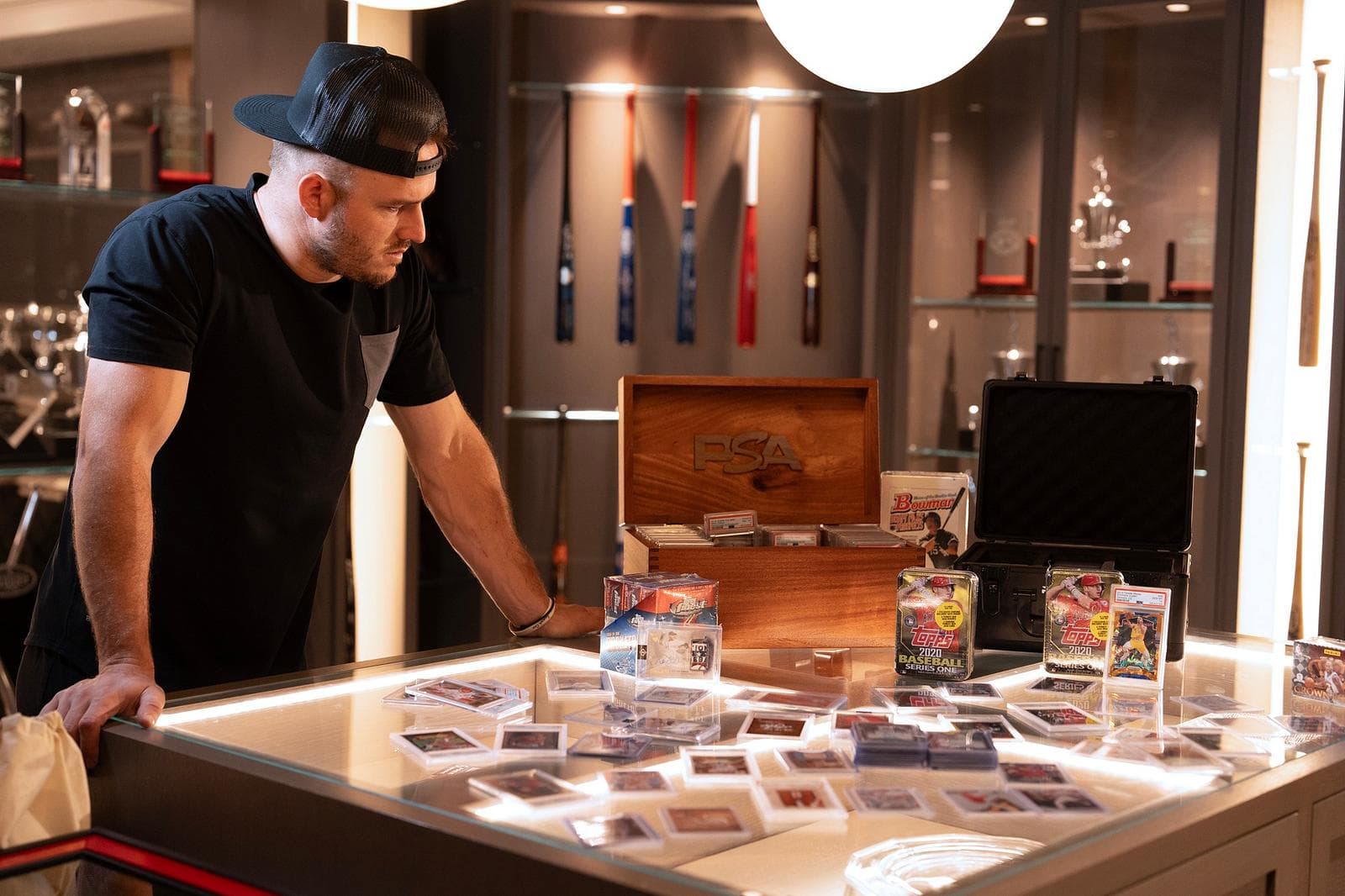 Mike Trout leans over a glass display case covered in sealed boxes and graded cards