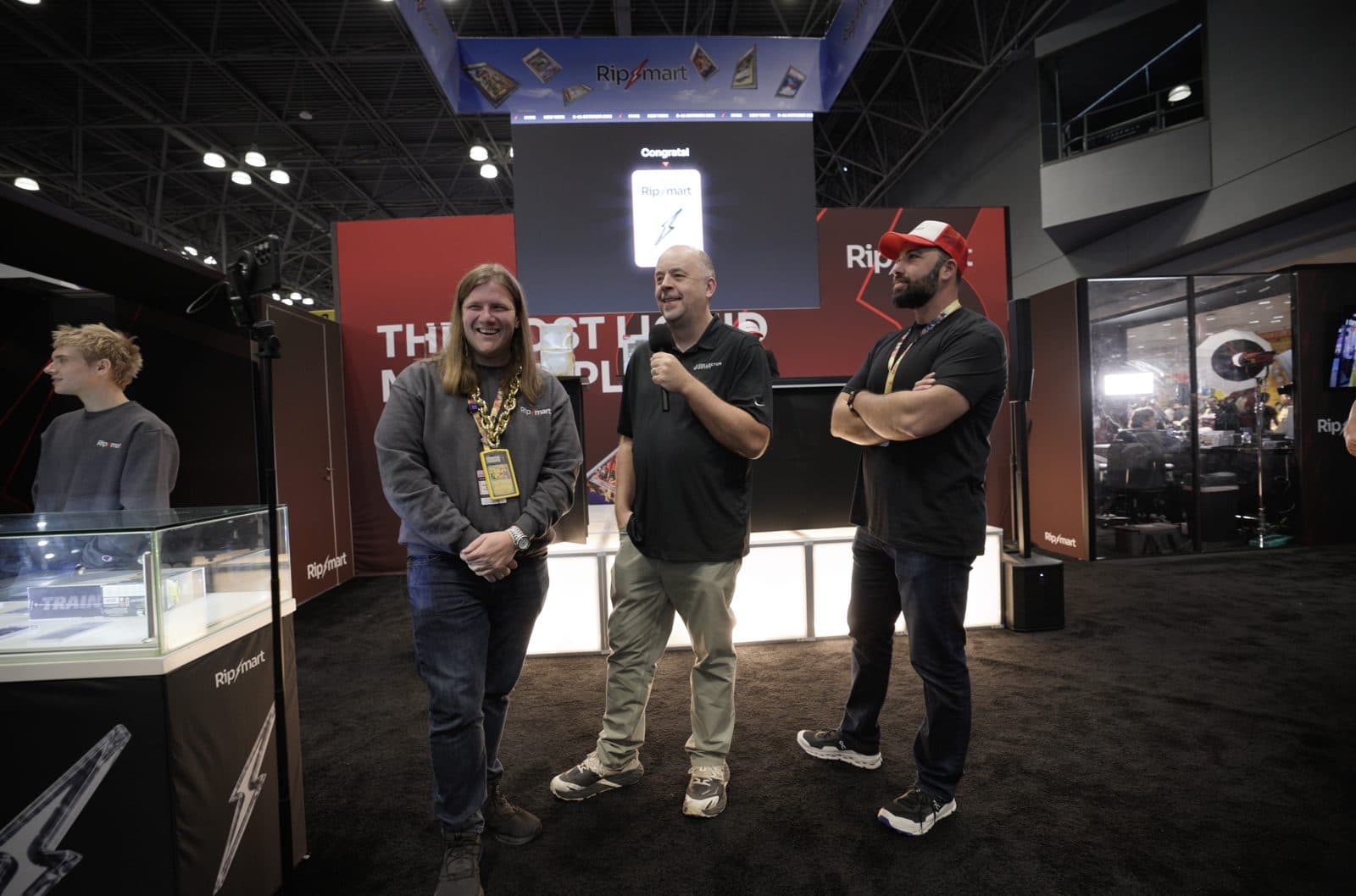 Three men stand on the Ripmart stage, one holding a microphone, under a screen that reads Congrats
