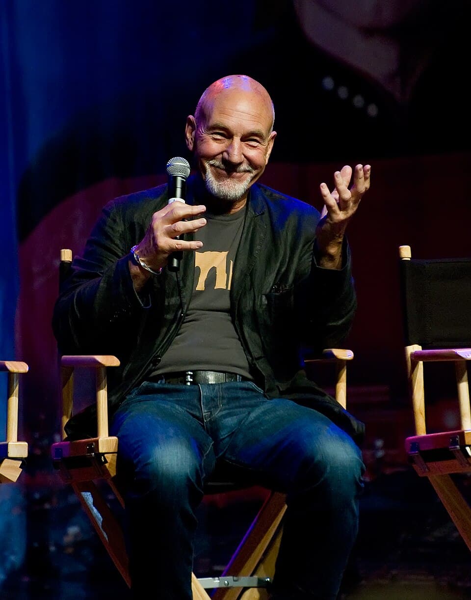 Patrick Stewart, smiling with a microphone in a director's chair, on stage at the Las Vegas Star Trek Convention in 2011.